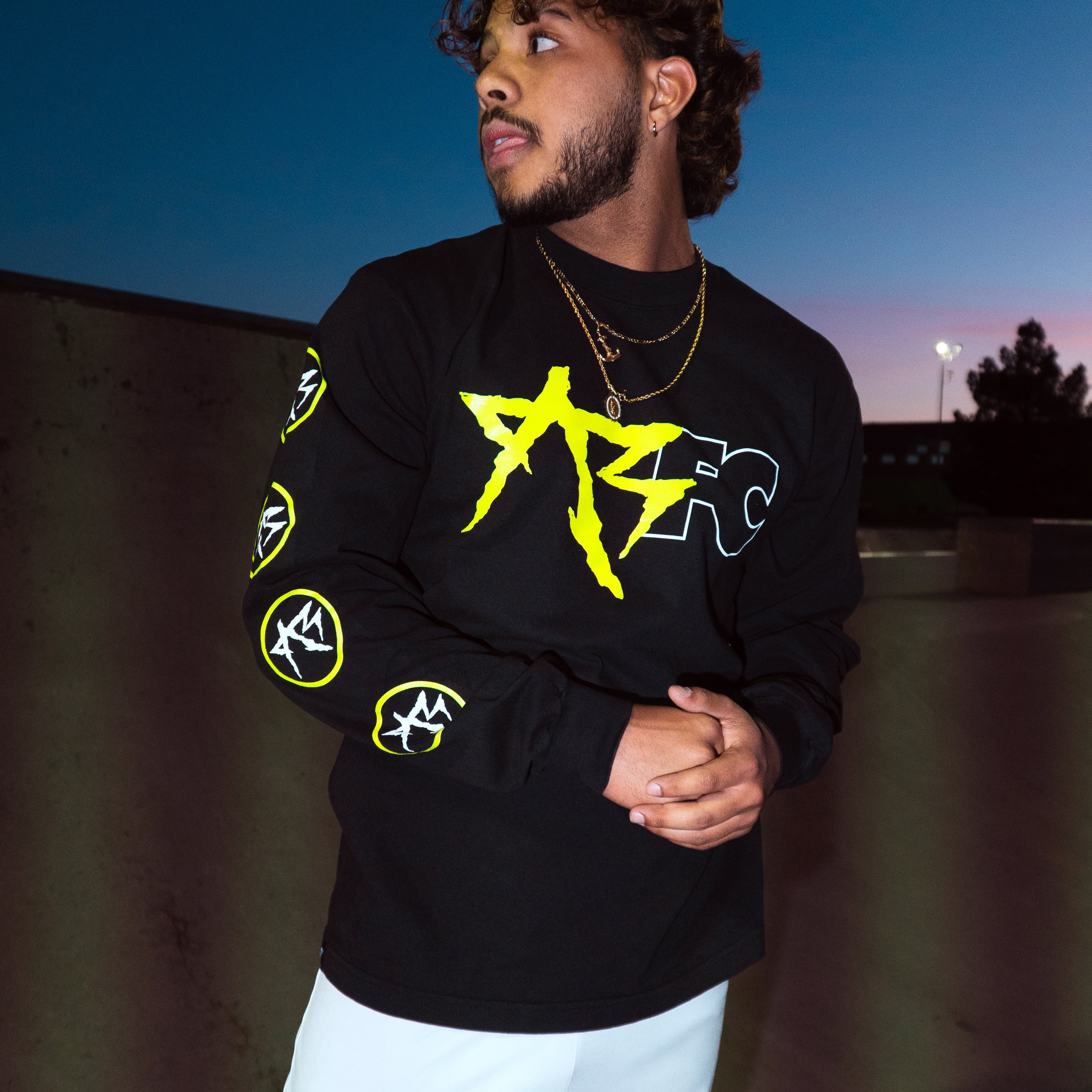 A man wearing a black long-sleeve graphic sweatshirt with a large yellow and white star design on the chest, paired with white shorts, standing outdoors against a twilight sky.