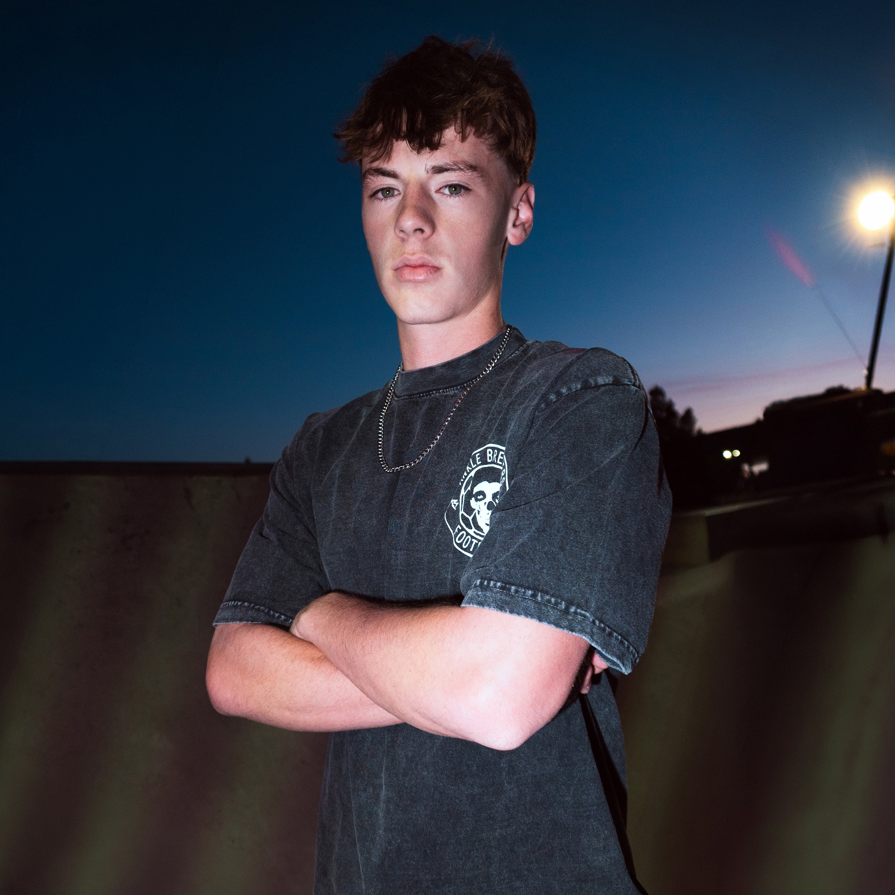 A young man with short brown hair stands outdoors at night with his arms crossed, wearing a faded black graphic t-shirt. The background features a dark blue sky, a distant street light, and a dark wall.