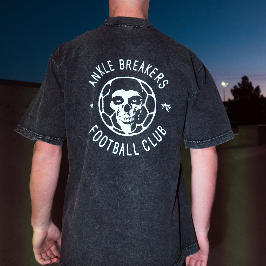 A man viewed from behind wearing a faded black t-shirt with a white graphic on the back that reads 'ANKLE BREAKERS FOOTBALL CLUB' around a skull and soccer ball design.