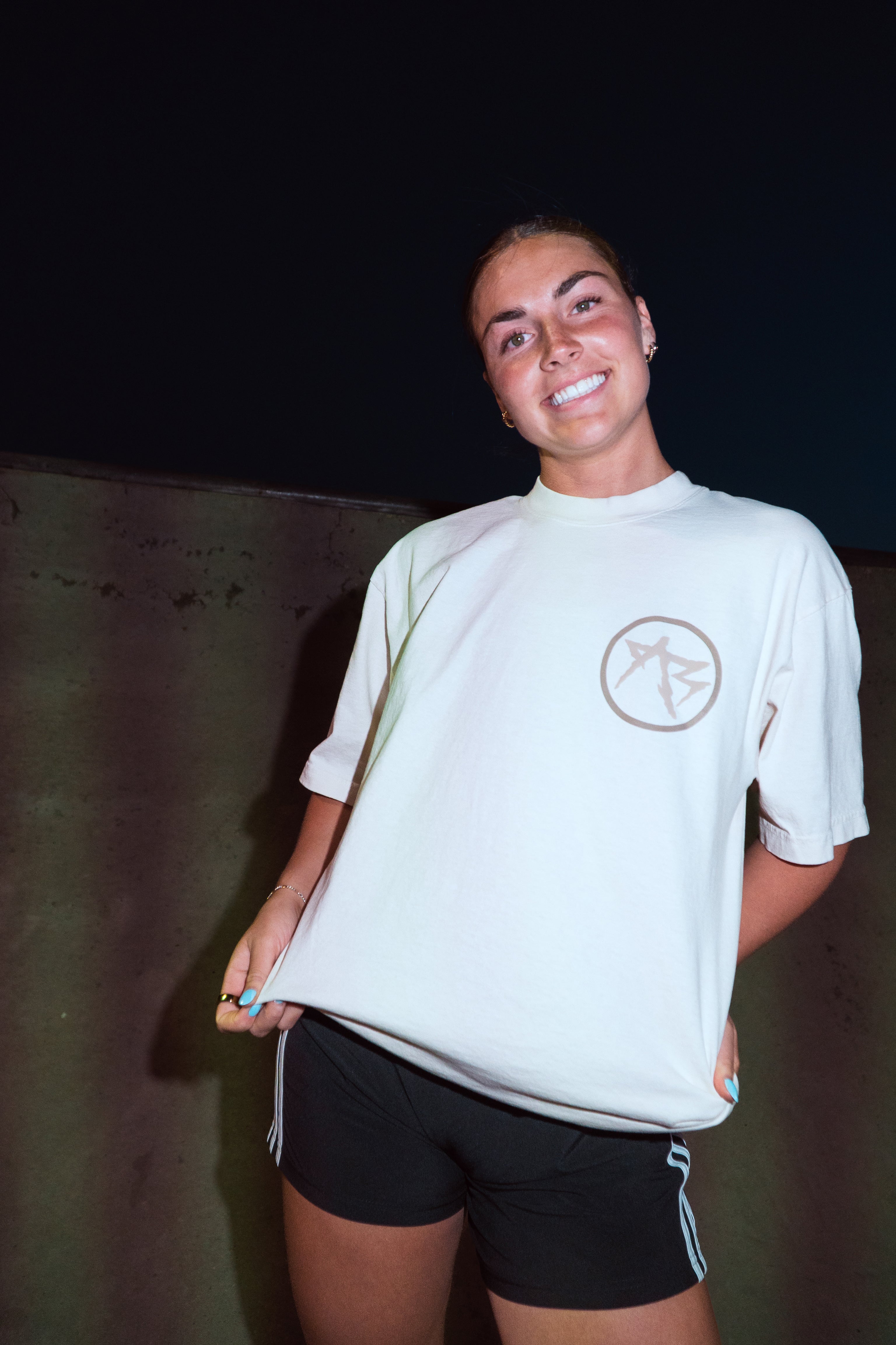 A woman wearing a white short-sleeve t-shirt with a brown circular logo on the left chest, paired with black athletic shorts, posing against a dark background.