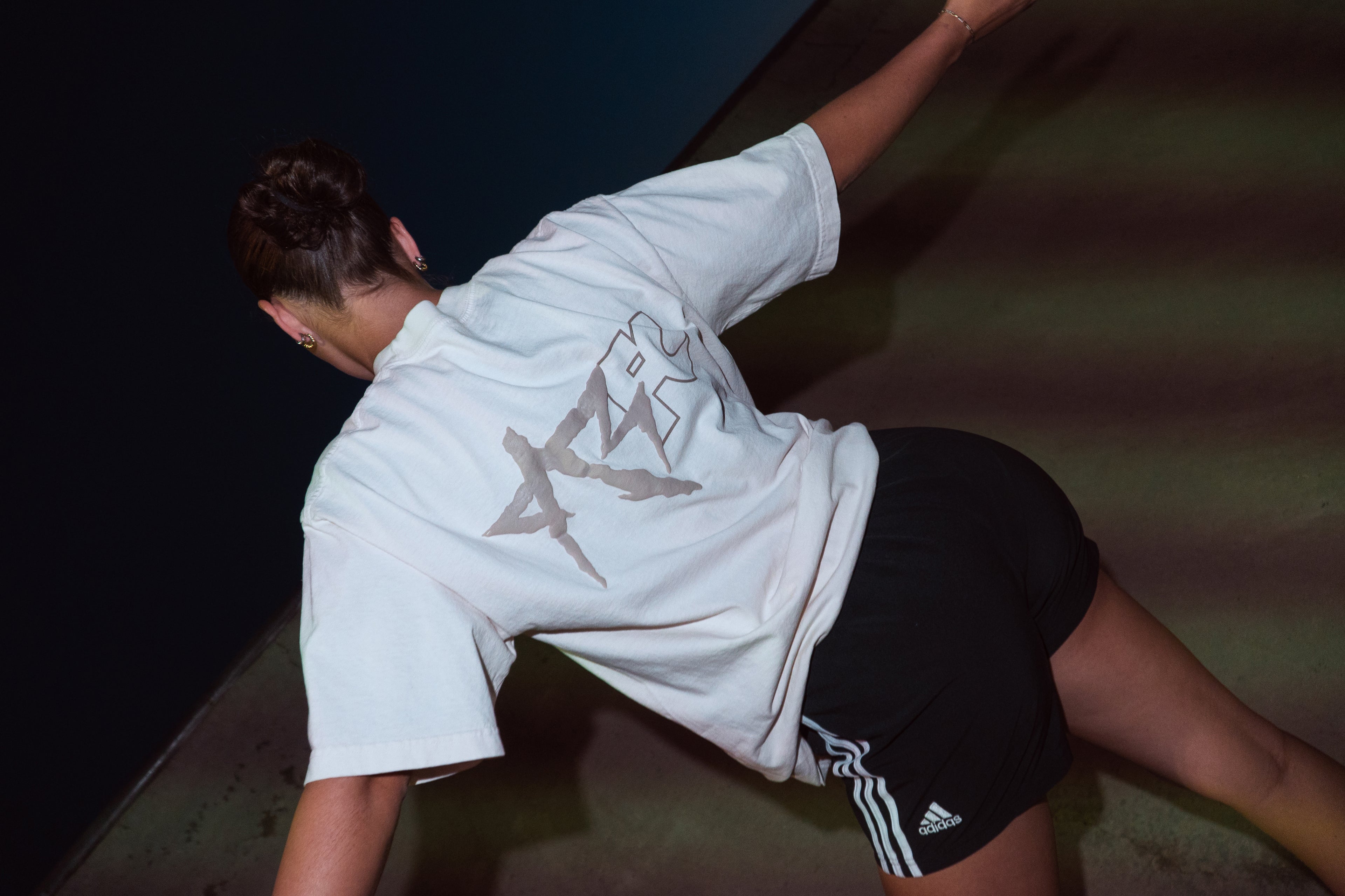 A person wearing an oversized white graphic T-shirt and black athletic shorts with white stripes, posed dynamically against a dark background.
