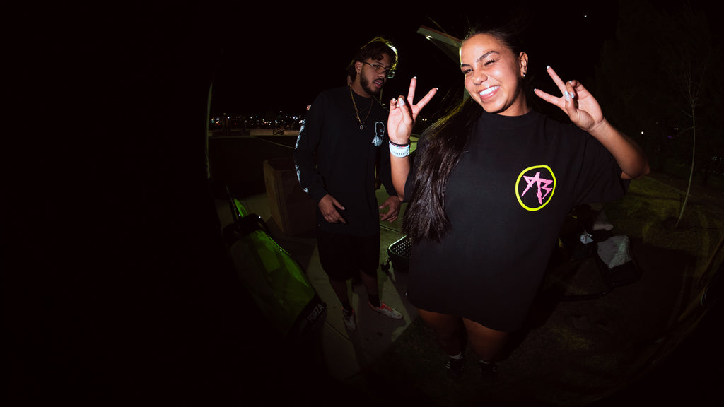 A woman posing at night wearing a black t-shirt with a yellow and pink circular graphic, making peace signs, with a man in a black shirt standing behind her.