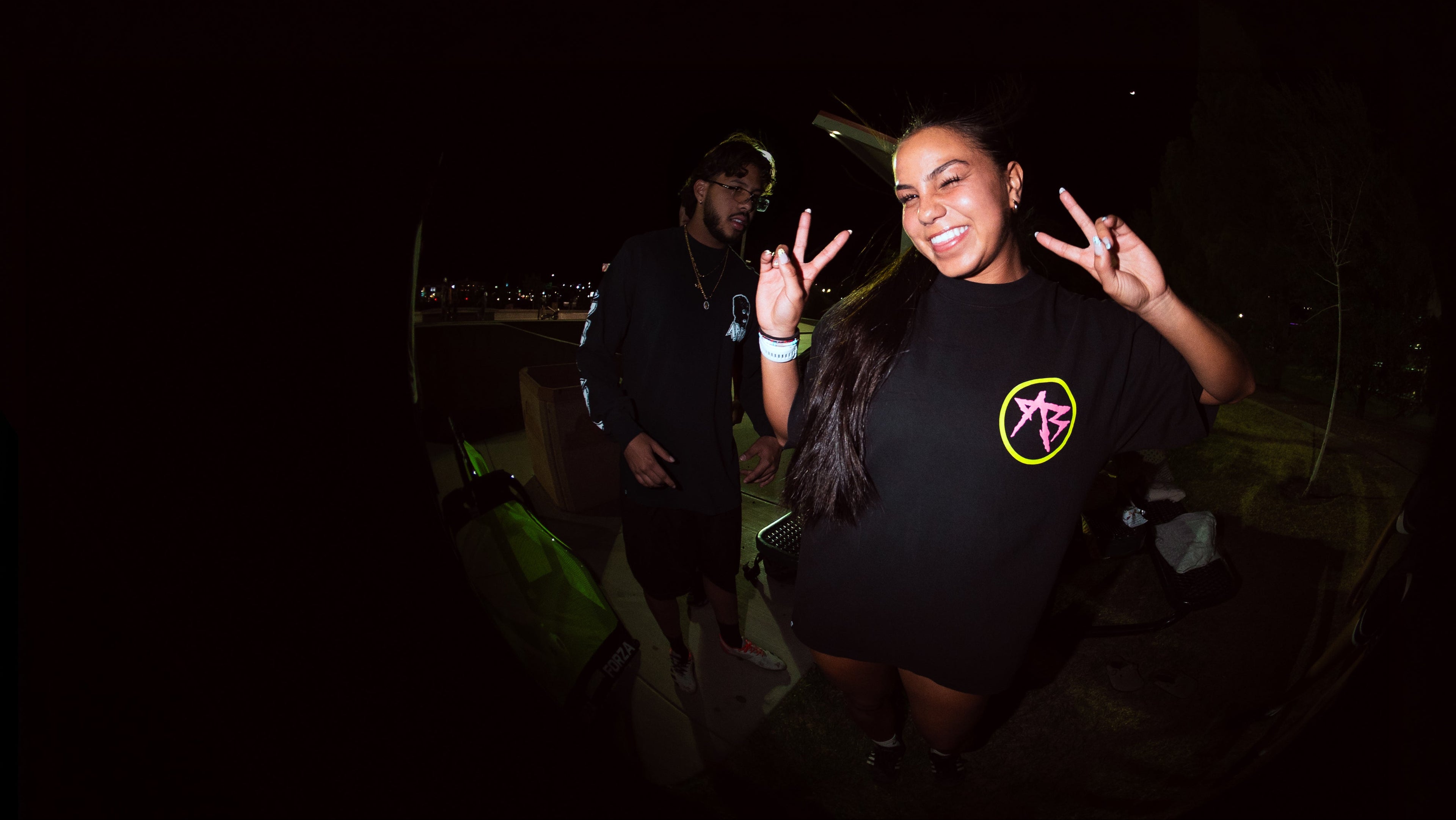 A woman posing outdoors at night wearing a black t-shirt with a pink and yellow circular graphic, making peace signs with both hands. A man is standing in the dark background.