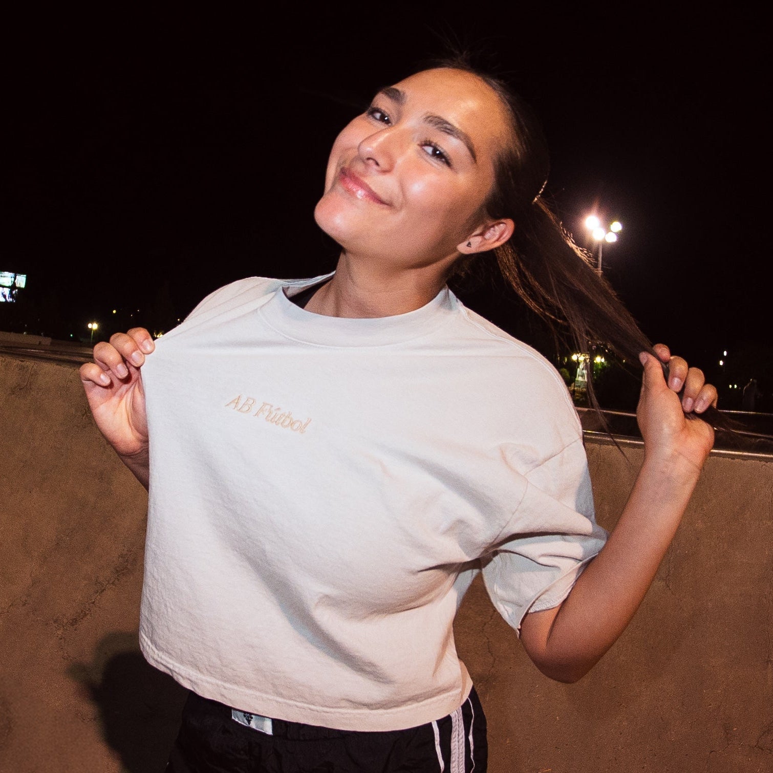 A woman posing in a skatepark at night under a fisheye lens, wearing a light gray t-shirt and black shorts.