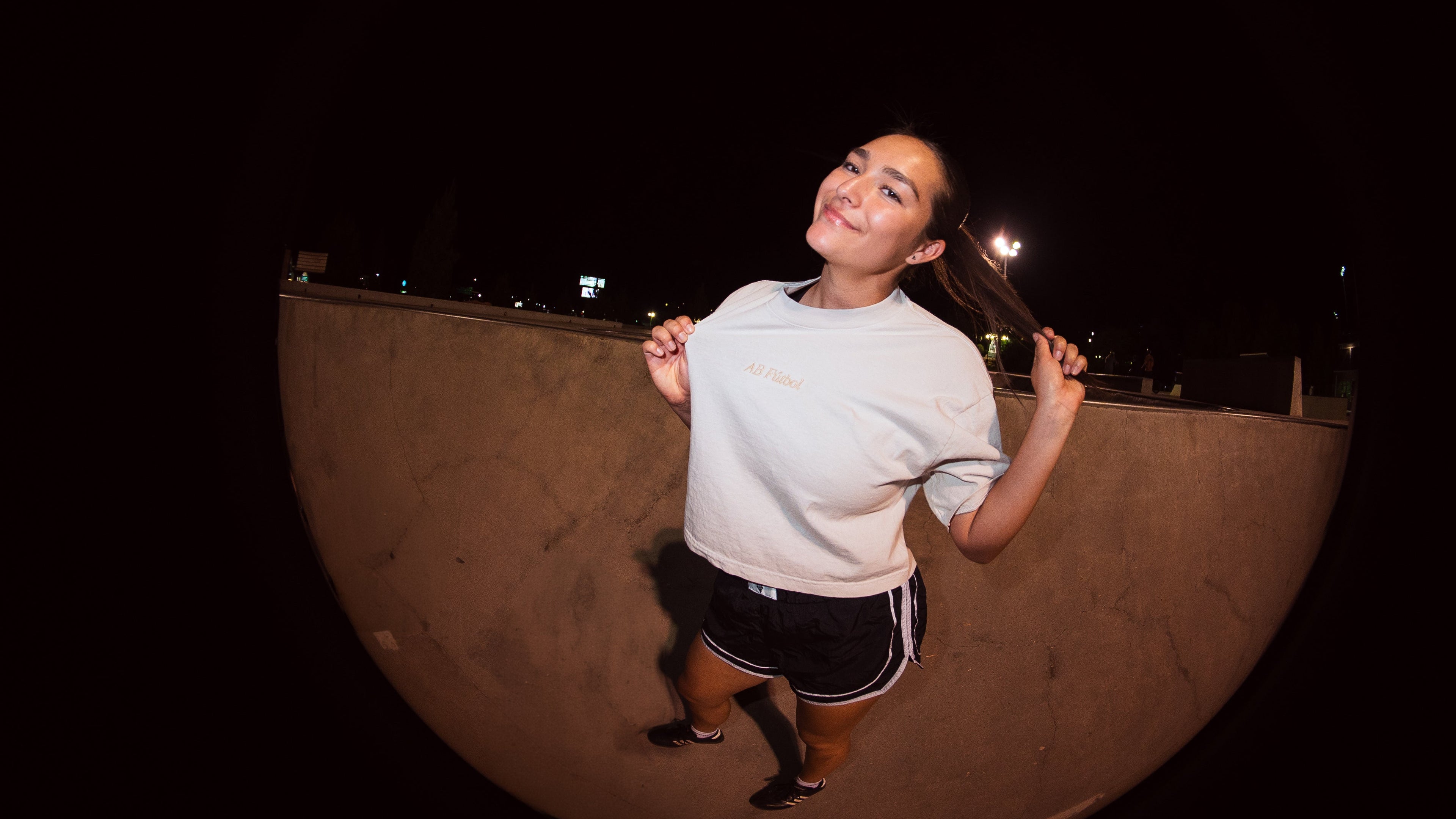 A woman posing in a skatepark at night under a fisheye lens, wearing a light gray t-shirt and black shorts.