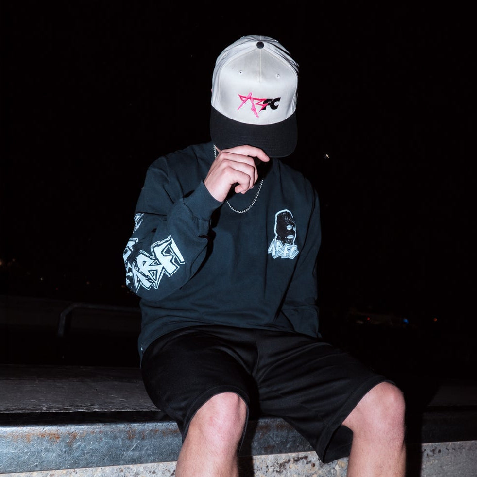 A person sitting on a dark outdoor ledge at night, wearing a light gray and black baseball cap, a black long-sleeve graphic shirt, and black shorts.